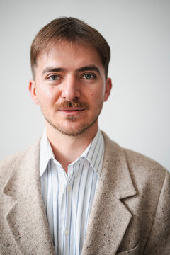 A young man with brown hair and a mustache wears a striped shirt and a tweed jacket, looking directly at the camera with a neutral expression.