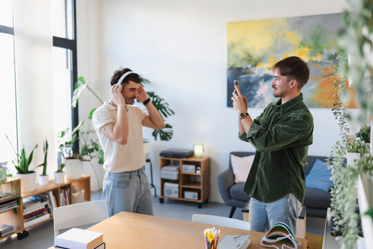 A man films his friend adjusting headphones in a bright, modern room with plants and artwork.
