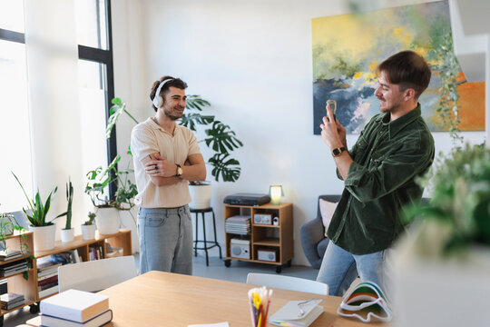 A man with headphones films his friend with a smartphone in a bright, modern room filled with plants and art.