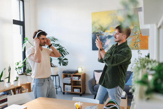 Two young men are in a bright, modern room. One man is putting on headphones while the other films him with a smartphone.