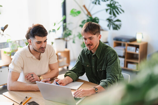 Two men collaborate on a project at a desk, using a laptop and notebook. They are engaged in discussion and planning, surrounded by plants in a bright, modern office space.