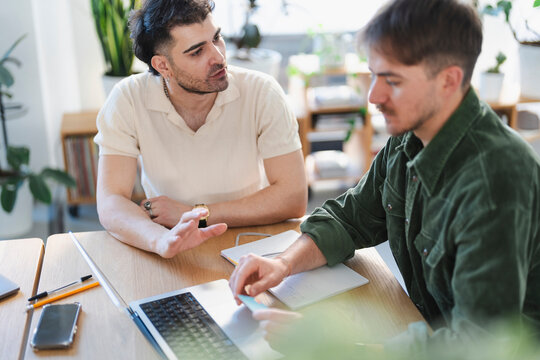 Two men collaborate at a table, one gesturing while the other holds a card near a laptop and notebook, suggesting a business meeting or study session.