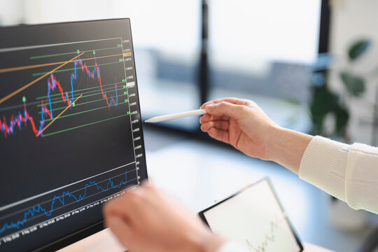 A person analyzes financial charts on a computer screen and a tablet, using a stylus to point at the data.