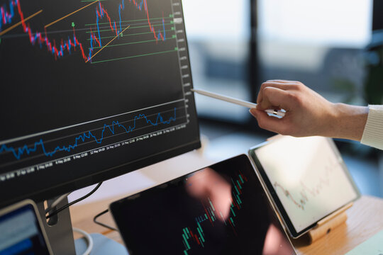 A person's hand holds a stylus, pointing at a financial chart on a computer monitor. Multiple tablets display similar trading data.