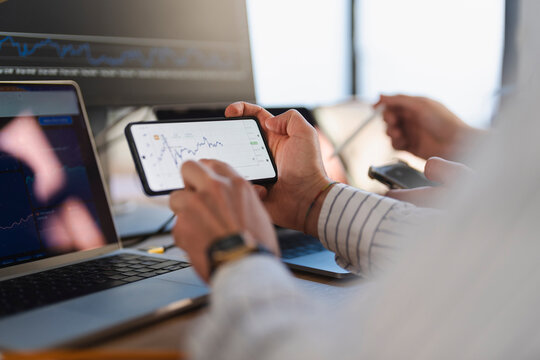 A person analyzes financial charts on a smartphone while another person uses a laptop, suggesting a collaborative trading or investment session.