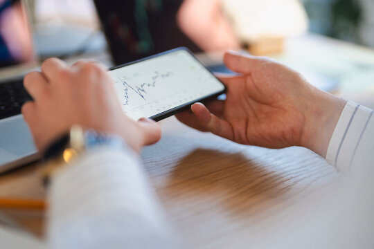A person holds a smartphone displaying a financial chart, with a laptop in the background, suggesting analysis of market trends.