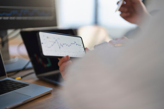 A person analyzes financial charts on a smartphone, with a laptop and computer monitor displaying market data in the background.