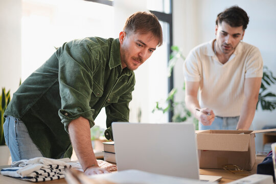 Two men are working together at a table, one looking at a laptop while the other prepares to write in a box.