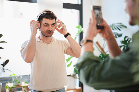 A man adjusts his headphones while another person films him with a smartphone, capturing a moment of personal enjoyment and digital sharing.