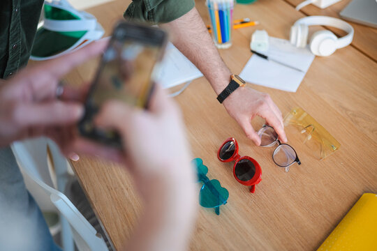 A person holds a phone while reaching for glasses on a wooden table with sunglasses and other desk items.