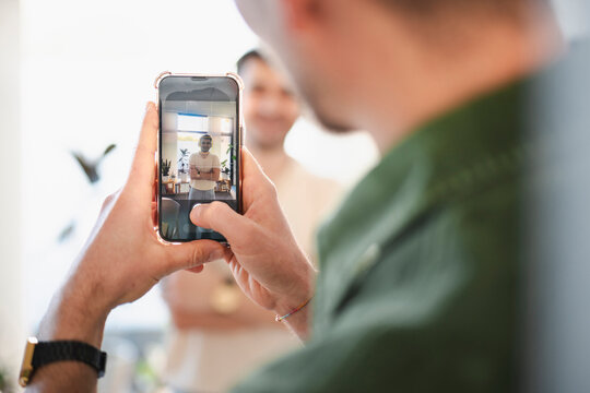 A person holds a smartphone, capturing a photo of another person standing with arms crossed in a bright room.