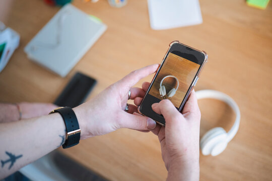 A person holds a smartphone, capturing a photo of white headphones on a wooden desk. Other items are visible in the background.