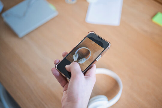 A person's hand holds a smartphone, capturing an image of white headphones on a wooden desk.