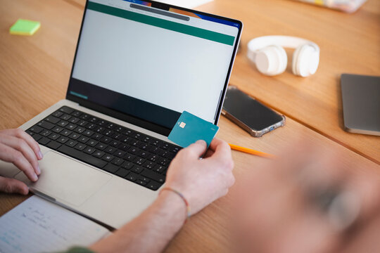 A person uses a credit card to make an online payment on a laptop, with a smartphone and headphones nearby on a wooden desk.