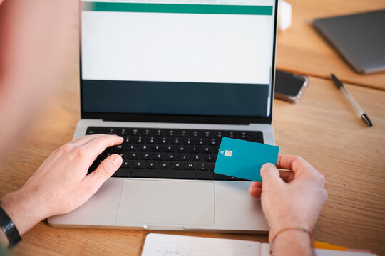 A person uses a debit card to make an online payment on a laptop, with a smartphone and notebook on the desk.