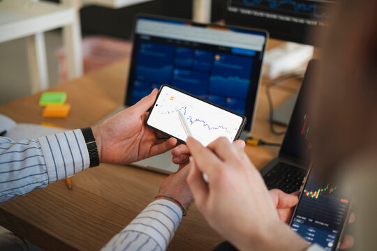Two people analyze financial charts on a smartphone and laptop, discussing market trends and investment opportunities in a modern office setting.