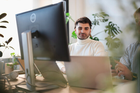 A man in a white turtleneck smiles while working at his desk with a laptop and computer.