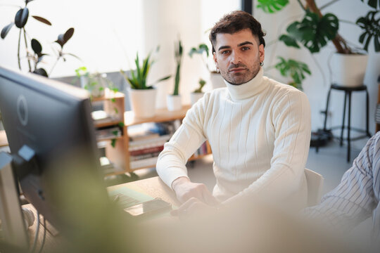 A man in a white turtleneck sits at a desk with a computer and phone, looking directly at the camera in a bright, plant-filled office.