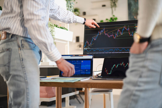 A person points to a financial chart on a large monitor, while another person stands nearby, reviewing data on a laptop.