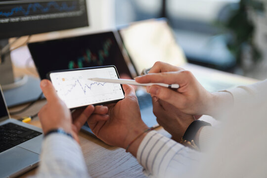 Two people analyze financial charts on a smartphone and laptop screens, discussing market trends and investment strategies.