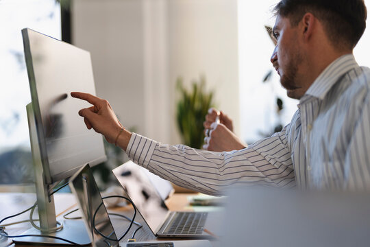 A man in a striped shirt points at a computer screen displaying charts, working at his desk with a laptop and other office supplies.