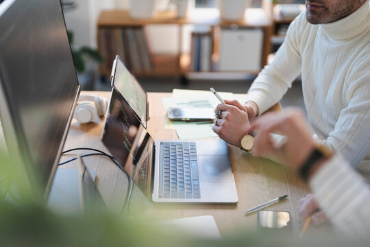 Two people collaborate at a desk, one holding a stylus and looking at a laptop screen displaying financial data, the other writing.