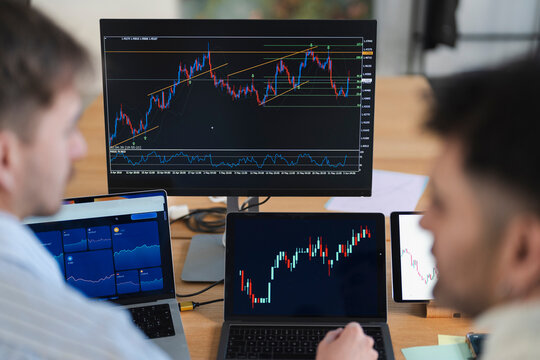 Two individuals analyze financial market charts on multiple screens, indicating a focus on trading and investment strategies.