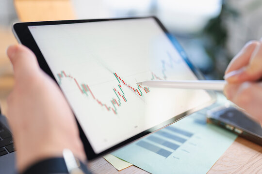 A person uses a stylus to analyze a financial chart on a tablet, with a smartphone and documents nearby.