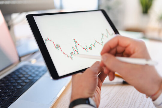 A person uses a stylus to interact with a financial chart displayed on a tablet, with a laptop in the background.