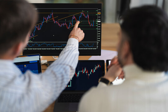 Two men analyze financial charts on multiple screens, one pointing to a complex graph indicating market trends and investment strategies.