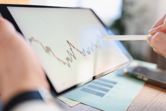 A person uses a stylus to analyze a financial chart on a tablet, with reports on the desk.