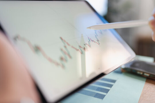 A person uses a stylus to analyze a financial chart on a tablet, with a phone and documents on a wooden desk.