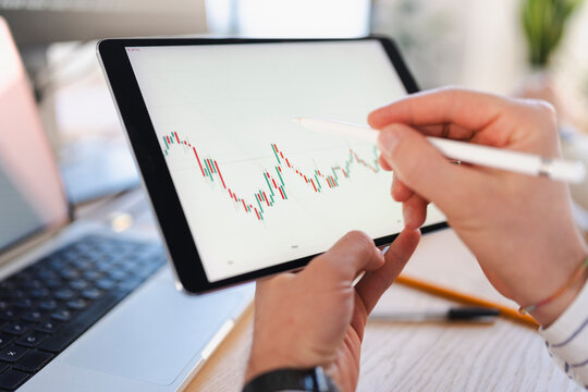 A person uses a stylus to interact with a financial chart displayed on a tablet, with a laptop in the background.