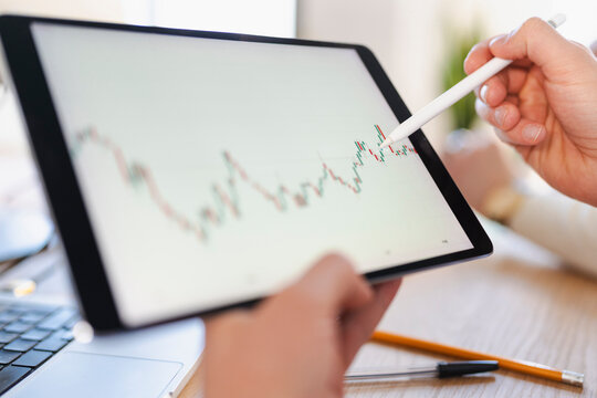 A person uses a stylus to interact with a financial chart displayed on a tablet, with a laptop and writing utensils on the desk.