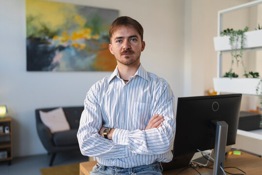 A young man with crossed arms sits in an office setting, looking directly at the camera. He wears a striped shirt and jeans, with a computer monitor and artwork in the background.