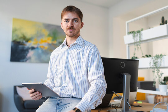 A man in a striped shirt sits on a desk in an office, holding a tablet. He looks directly at the camera.