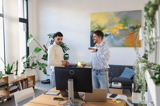 Two men collaborate in a modern office space, discussing documents and working on a laptop.