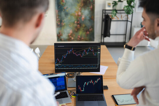 Two men analyze financial charts on multiple screens, discussing market trends and investment strategies in a modern office setting.
