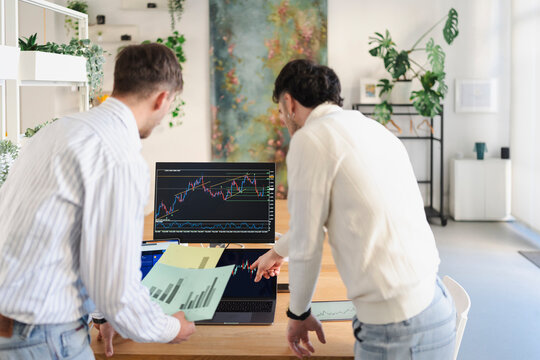 Two men analyze financial charts on multiple screens, discussing market trends and data in a modern office setting.