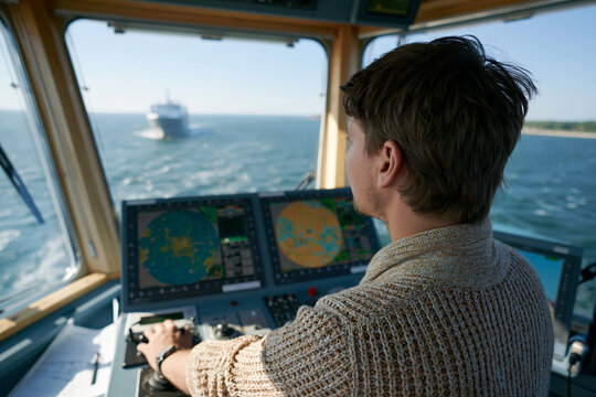 Young captain controls vessel at sea. Focus on navigation screens and steering controls. Sunny day, calm blue waters surround ship. Focused expression shows professional control