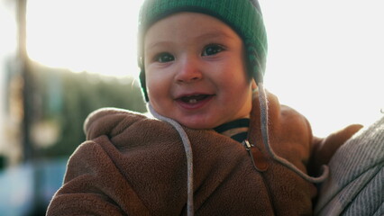 Baby in bear teddy fleece coat and green beanie hat smiling directly at camera outdoors, cheerful toddler showing first teeth and enjoying winter season family moment © Marco