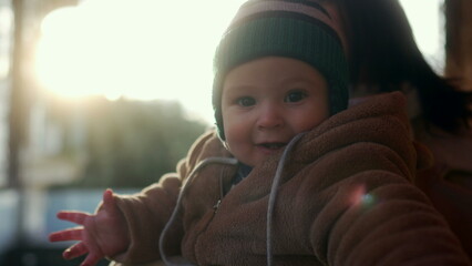 Baby in bear teddy fleece coat and striped beanie hat bringing hands to mouth while mother in beige coat holds child outdoors, cozy and tender family moment in cold season sunlight © Marco