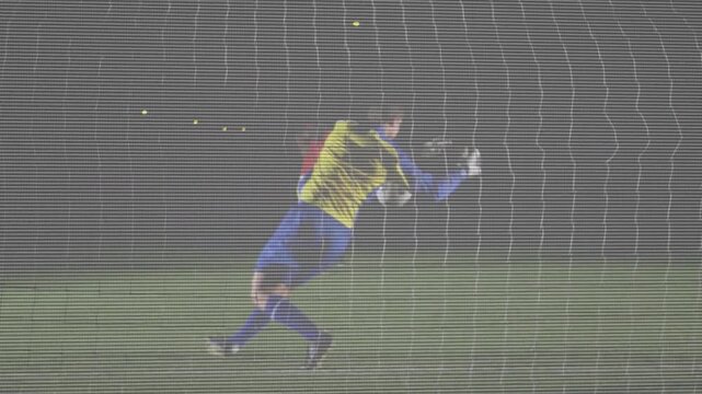 Goalkeeper diving after attacker striking ball, protecting soccer goal through net under lights