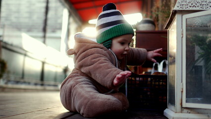 Baby in bear teddy fleece coat and striped beanie hat outdoors reaching toward lantern, toddler exploring environment with curiosity during winter afternoon light © Marco