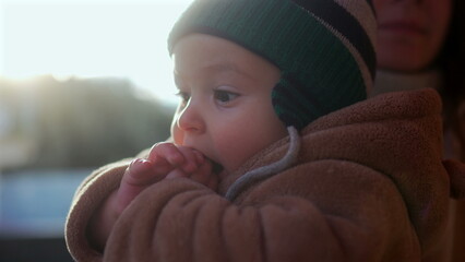 Baby in bear teddy fleece coat and striped green beanie hat with curious expression outdoors, infant looking ahead while sitting in parent’s arms during winter afternoon © Marco