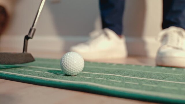 Slow motion close up putting a golf ball on an indoor putting trainer mat with white sneakers planted and a putter face approaching. Golf training videos and product ads.