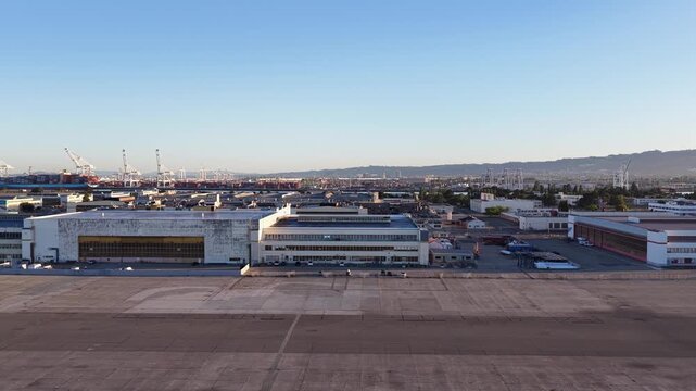Aerial view flying sideways over the former Alameda Naval Air Station, showing the historic hangars and the port of Oakland cranes across the water under a clear blue sky at sunrise