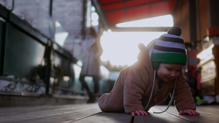 Baby crawling outdoors in bear teddy fleece coat and striped beanie hat with mother in beige coat walking behind, toddler exploring wooden floor during cold season day © Marco