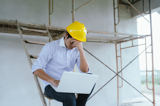Stressed engineer wearing a safety helmet using a laptop at a construction site, representing project pressure, workplace problems, and construction management.