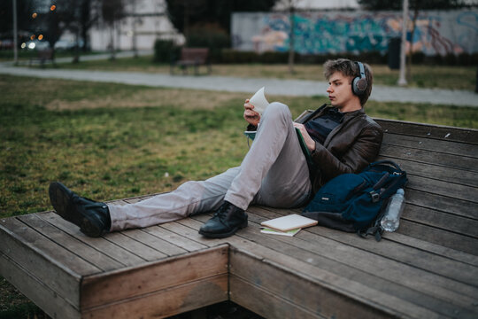 A high school student wearing headphones reads a book while reclining on a wooden park bench. The young student relaxes outdoors with a backpack and notebooks beside him.